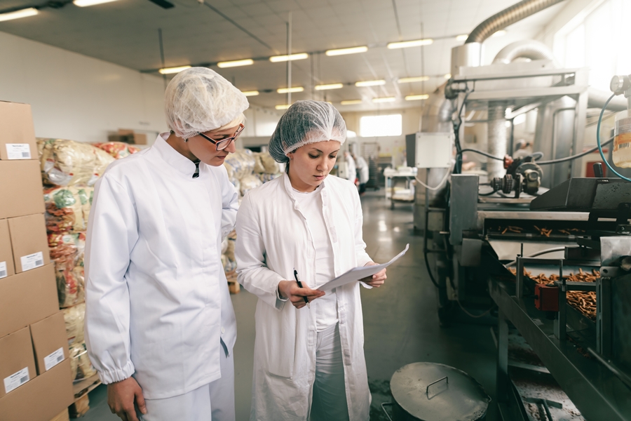 Two quality professionals in white sterile uniforms checking quality of salt sticks while standing in food factory.