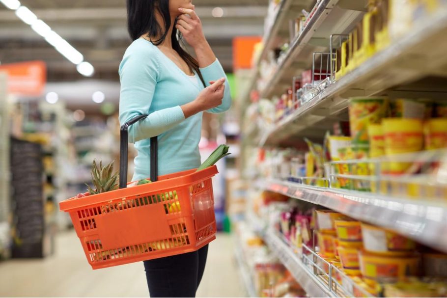 A woman holds a basket in a grocery store and looks at a shelf of items