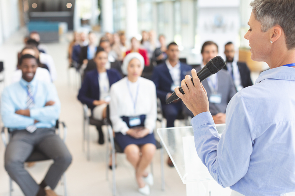 Rear View Of Caucasian Female Speaker Speaks With Microphone To